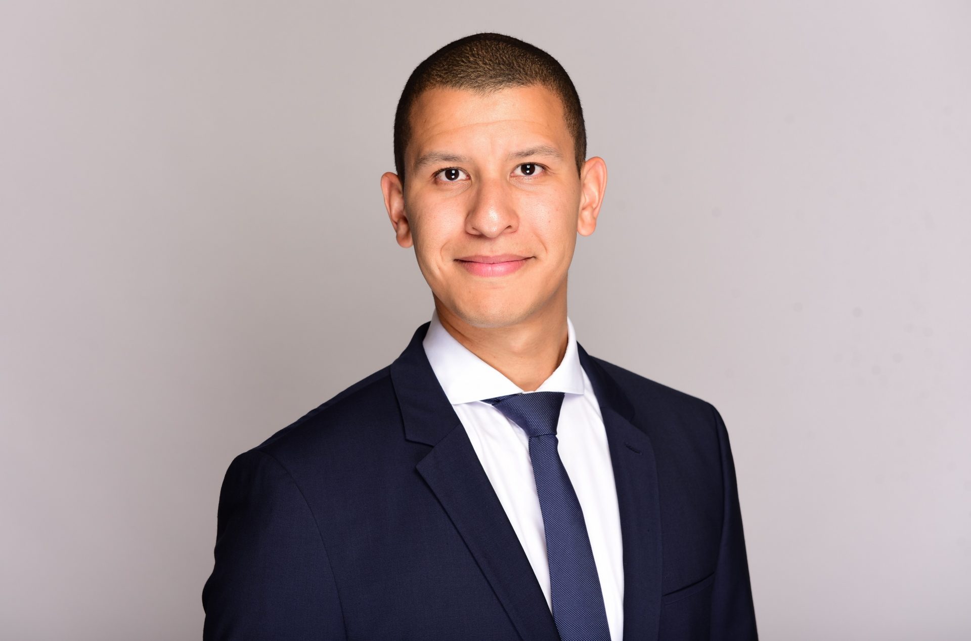 The image shows a person wearing a dark suit with a white shirt and a tie. The background is plain and gray.