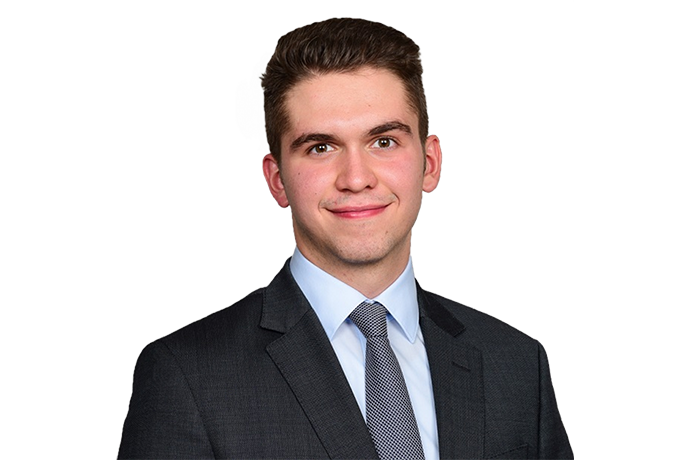 The image shows a person wearing a dark suit with a white shirt and a tie on a black background.
