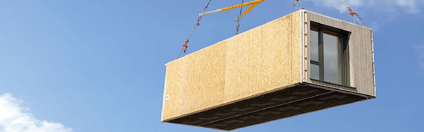 The image shows a prefabricated building module being lifted by a crane in an outdoor setting. The module resembles a container-like structure with wooden paneling on the exterior and glass windows. This process is part of modular construction, where individual sections or units of a building are manufactured off-site and then transported to the desired location for assembly.