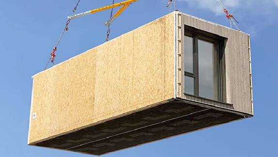 The image shows a prefabricated modular building unit being lifted by a crane. Key features of the unit include:

1. **Structure Material:** The main body of the unit appears to be constructed from a form of engineered wood or composite materials, visible through the textured wooden facade.

2. **Design:** The unit features a large glass window, suggesting that it is designed for residential or office use.

3. **Lifting Mechanism:** Steel cables are attached to the corners of the unit, indicating it is being hoisted by industrial machinery, which is typically used for transporting and positioning prefabricated buildings.

4. **Environment:** The clear blue sky provides a backdrop, emphasizing that the unit is being lifted outdoors, likely as part of a construction or installation process.

5. **Purpose:** Such modular units are often used for quick assembly of structures like temporary housing, mobile offices, or tiny homes.

This image showcases modern construction techniques focused on efficiency and sustainability, leveraging prefabricated components that can easily be transported and assembled on-site.