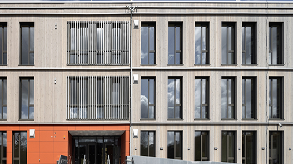 The image depicts a modern building exterior. The structure has a clean, minimalist design characterized by:
- **Vertical Windows**: The facade comprises multiple windows arranged in vertical lines. Each window has a square or rectangular shape, appearing uniform across the building face.
- **Concrete Panels**: The exterior material seems to be concrete or a similar composite, providing a sleek, smooth look.
- **Central Entrance**: A primary entryway section is visible at the center, distinguished by an orange panel that adds contrast to the surrounding neutral tones. The entrance is recessed, providing a sheltered space.
- **Metal Bars or Grills**: Some parts of the facade have metallic bars or grills over certain sections, possibly for aesthetic balance or functional purposes like sun shading or security.
- **Reflective Glass**: The windows appear to be made of reflective glass, showcasing sky reflections, which accentuate the modern architectural feel.
- **Urban Setting**: The surrounding context appears urban, evidenced by the building's contemporary design suited for an office, institutional, or residential application.