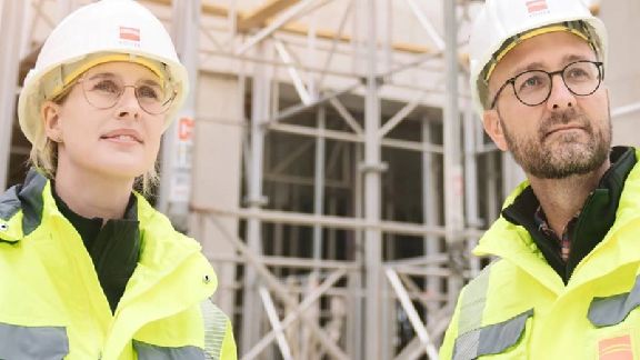Two individuals wearing construction safety gear, including hard hats and high-visibility jackets, standing in front of scaffolding at a construction site.