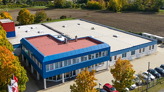 The image depicts a medium-sized building in an aerial perspective. The building has a distinct architectural design featuring a flat roof and rectangular shape with blue accents and white facade. The structure appears to be an office or a small commercial building, characterized by its modern design and clean lines.

Features:

1. **Flat Roof:**
   - The building's roof is flat, with some equipment or installations visible, likely related to climate control systems.

2. **Color Scheme:**
   - The building has a blue perimeter framing its walls and windows, standing out against the primary white facade.

3. **Surrounding Area:**
   - The building is located in a landscaped area with trees that are showcasing fall colors, suggesting an autumn season.
   - There are agricultural fields in the background, indicating a more rural or suburban setting.

4. **Parking Lot:**
   - In front of the building, there is a parking lot visible, with several cars parked, indicating occupancy.

5. **Flags:**
   - Near the entrance or roadside of the building, there are flags likely representing branding or organizational presence.

6. **Windows:**
   - Large windows are visible on the blue-colored section of the building, allowing for significant natural light.

Overall, the image depicts a commercial or office environment set within a suburban or rural landscape, highlighting the contrast between modern infrastructure and natural surroundings.