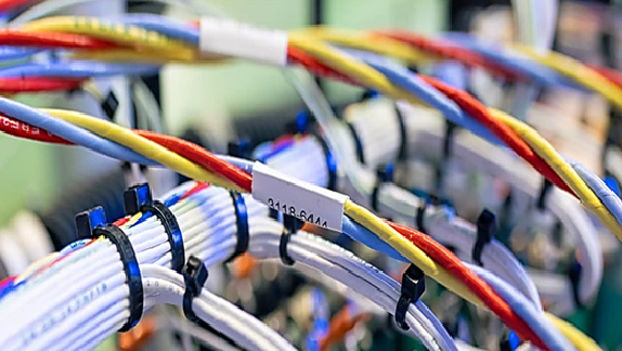 The image depicts a close-up view of a collection of wires or cables that are part of an organized wiring system. The cables are bound together using cable ties or clips to keep them neatly arranged. The cables appear to be labeled, suggesting they might be part of a complex electronic or networking system. The presence of red, yellow, blue, and white colored cables indicates that different types of cables are possibly being used, each with a specific function or signal type. This arrangement is typical within an electrical or computer network installation, such as in a data center or server room.