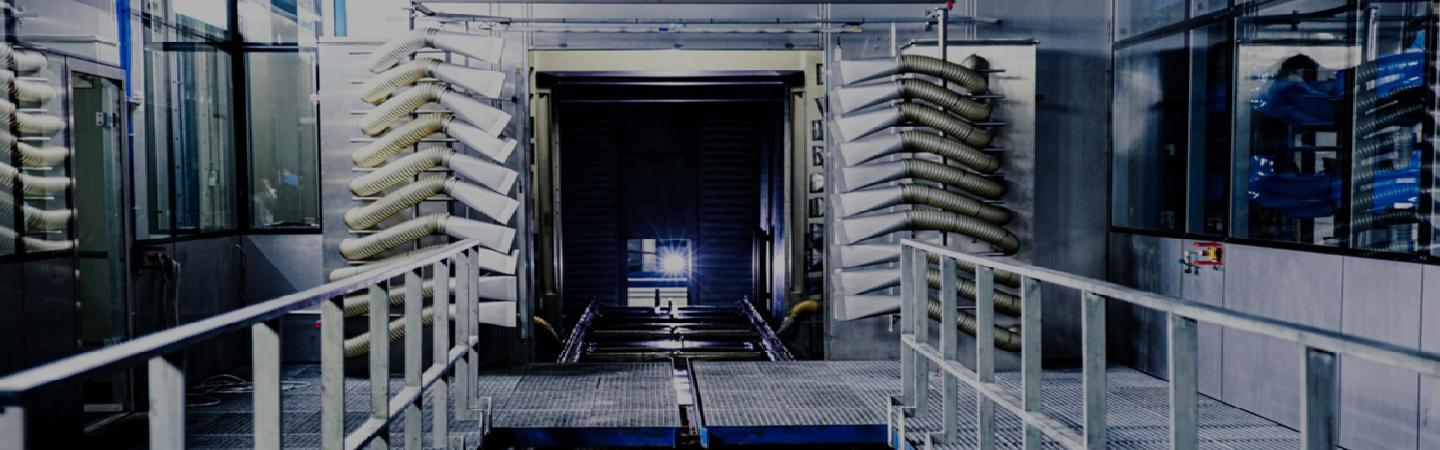 The image appears to depict an industrial setting, likely part of a manufacturing or processing plant. Key details observable include:
1. **Infrastructure:**
- The presence of metallic railings and platforms that suggest a walkway or operational area for workers.
- Walls and floor with a sterile, metallic design—indicative of environments that require cleanliness, such as food processing or pharmaceutical manufacturing.
2. **Equipment:**
- To the left and right of the central part of the image are coiled metallic or plastic pipes/hose systems, which may be used for transporting liquids or gases in industrial processes.
- Machinery or processing units that seem to be part of a conveyor system or tunnel, which could imply the transit of products or materials through a specific process.
3. **Lighting:**
- Bright, highly focused lights illuminating the area to enhance visibility, which is crucial for operational safety and precision.
4. **Environment:**
- A sense of order and structure that is typical of manufacturing plants, where materials are handled in a controlled environment.
This area could potentially be used for processes related to:
- **Automobile Manufacturing:** Parts could be coated, painted, or treated.
- **Food Processing:** Sterilization or packaging sections.
- **Material Testing:** Examination or treatment of industrial products.
Overall, it's an industrial image suggesting a highly organized, process-driven environment.