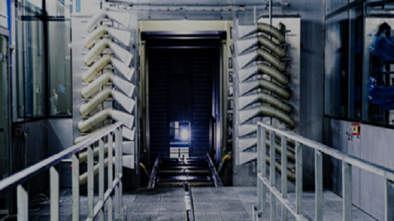The image depicts a highly industrial setting, possibly inside a factory or processing plant. Key features of the image include:

1. **Mechanical Equipment:** The presence of several tubes or pipes that are part of a larger machinery setup. They are seen vertically aligned and adjacent to the central opening.

2. **Metal Railings:** Metal railings can be seen to the sides of what appears to be a pathway leading to the central passageway or tunnel.

3. **Central Passageway:** There is a path or corridor with tracks, presumably allowing movement of equipment or materials.

4. **Lighting:** An illuminated spot or light source is visible in the center, possibly used for guiding or providing clarity down the passage.

5. **Glass Panels:** To each side of the equipment setup, glass panels can be observed, likely part of a ventilation or containment system.

6. **Industrial Environment:** The overall grey metallic tone affords the image a cold, mechanical feel indicative of an industrial environment.

This setting is typical of manufacturing plants, refining stations, or any facilities involved in large-scale production or processing activities.