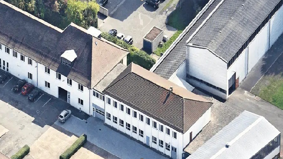 The image appears to display an aerial view of a facility or industrial complex.
### Key Observations:
1. **Buildings:**
- The main focus is on a few buildings that form part of what might be seen as a compound or campus.
- The architecture suggests these may be factory or warehouse buildings, characterized by their rectangular shape and large roof areas.
2. **Roof Types:**
- Different parts of the building complex have distinct roofing styles that might include flat, sloped, or ridged roofs.
3. **Parking and Vehicles:**
- Visible vehicles are parked in designated areas around the building, indicating either an office building or a factory where employees work.
4. **Surrounding Area:**
- The premises seem to have paved areas and possibly include transport paths or small road access.
- Greenery and landscaping appear to be minimal, suited for industrial spaces.
5. **Additional Structures:**
- There seems to be smaller subsidiary structures which might be storage areas or utility facilities.
6. **Contextual Clues:**
- The image portrays an organized layout typically found in business districts or manufacturing zones.
Overall, this image likely represents a commercial or industrial property, designed for logistics, manufacturing, distribution, or administrative functions.