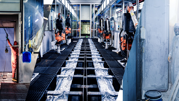 The image depicts a highly industrial setting, possibly a car factory or manufacturing plant. Key elements in the image include:
1. **Robotic Arms:**
- Multiple robotic arms are visible on either side of the central walkway. These are likely used for various automated processes such as assembly, welding, painting, or inspection.
2. **Protective Coverings:**
- There are silver protective coverings on the walkway or the section beneath the walkway, possibly serving as part of the process equipment or conveyor system.
3. **Workers in Safety Gear:**
- At least two individuals can be seen in the image, dressed in safety gear such as suits, masks, and gloves. These personnel may be responsible for manual inspection, oversight, or intervention when needed.
4. **Industrial Infrastructure:**
- The facility features columns and structure typically associated with large-scale manufacturing environments, possibly indicating production of automotive parts or heavy machinery.
5. **Bright Interior Lighting:**
- The setting is well-lit, providing clear visibility of the automated machinery and the tasks being performed.
6. **Clean Operation Zone:**
- The area is maintained clean and organized. It reflects an environment where precision and cleanliness are prioritized, which is common in automotive or mechanical processes.
